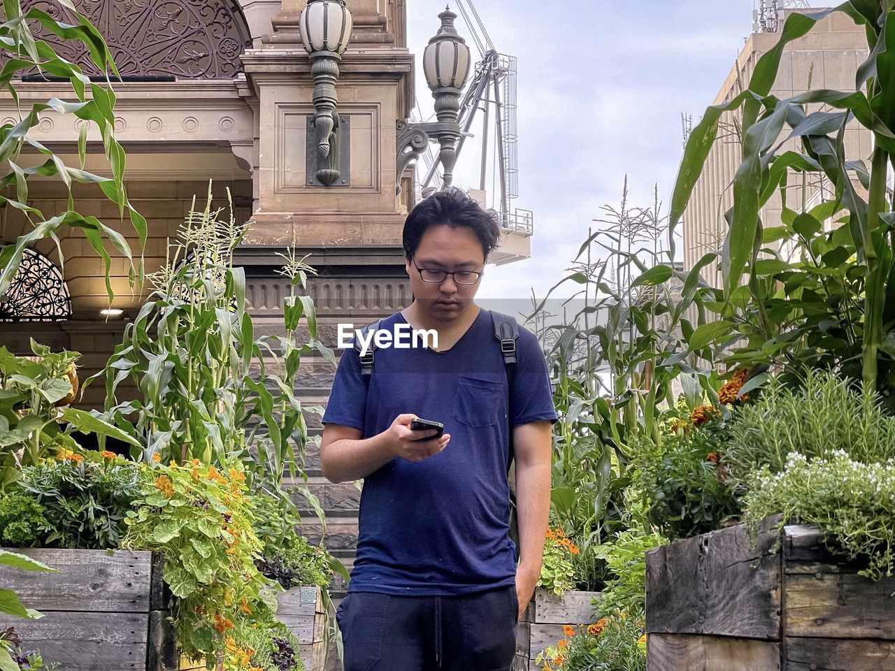 Portrait of young man using mobile phone in between flowering plants against buildings in the city.