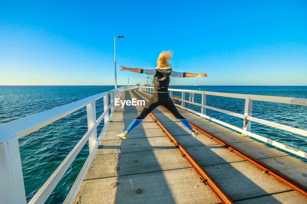 Full length of excited woman jumping on pier by sea against sky