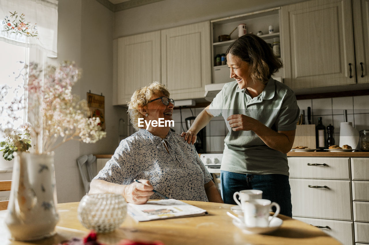 Happy female caregiver with elderly woman sitting near dining table in kitchen at home