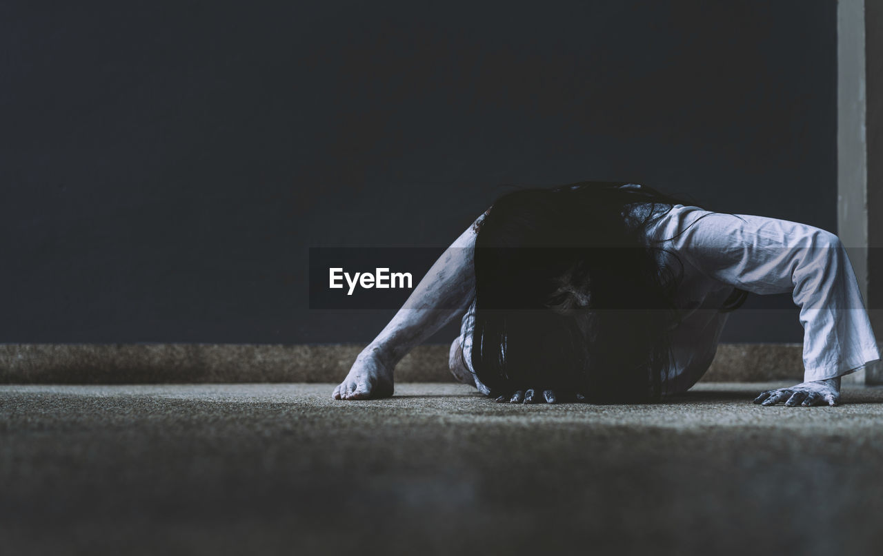 Spooky young woman crouching on floor against wall