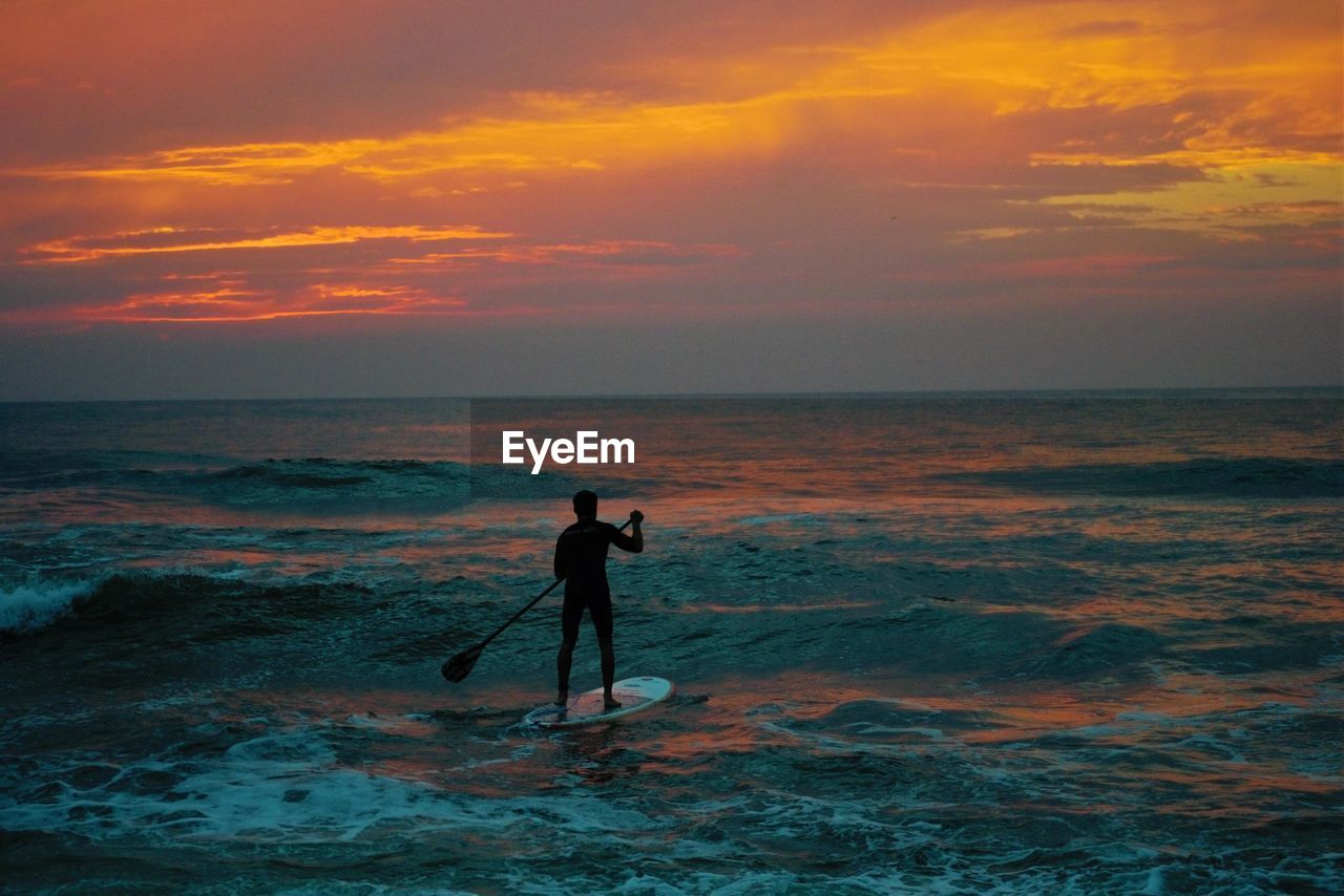 Silhouette man paddleboarding in sea against sky during sunset