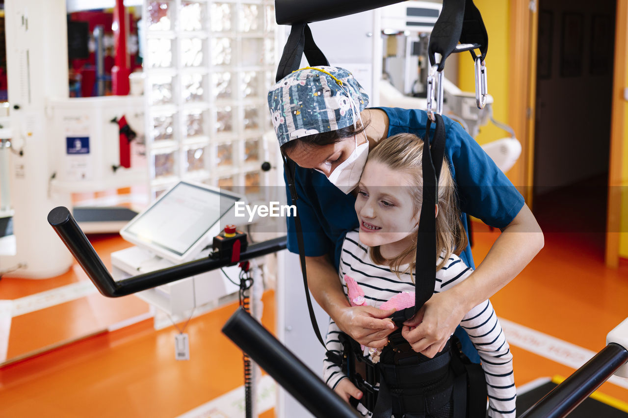 Professional medical specialist helping little girl with angelman syndrome and fixing straps while preparing for physiotherapy training on modern equipment in rehabilitation center