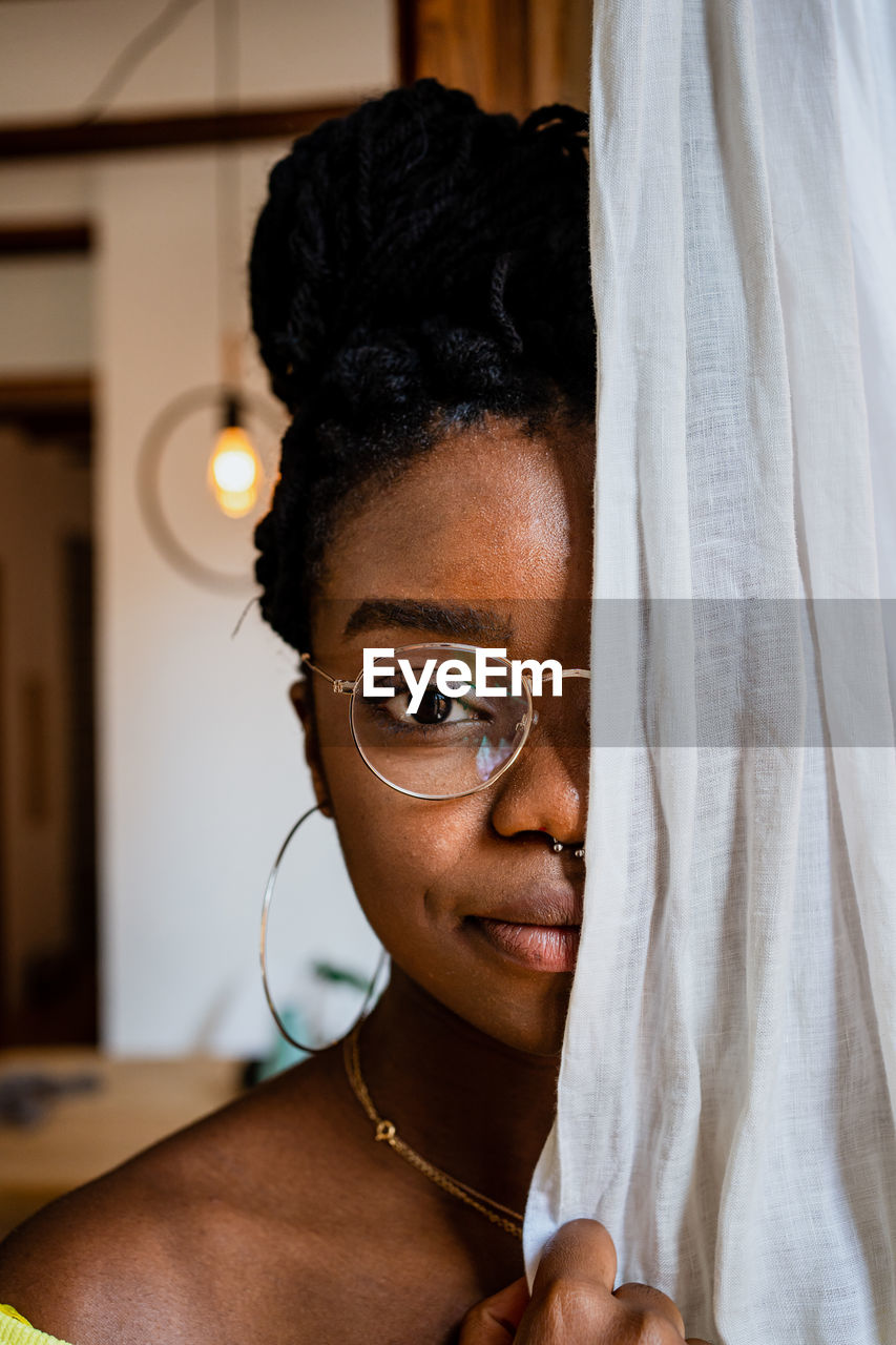 Pensive young african american woman in trendy eyeglasses covering half of face with curtain and looking at camera at home