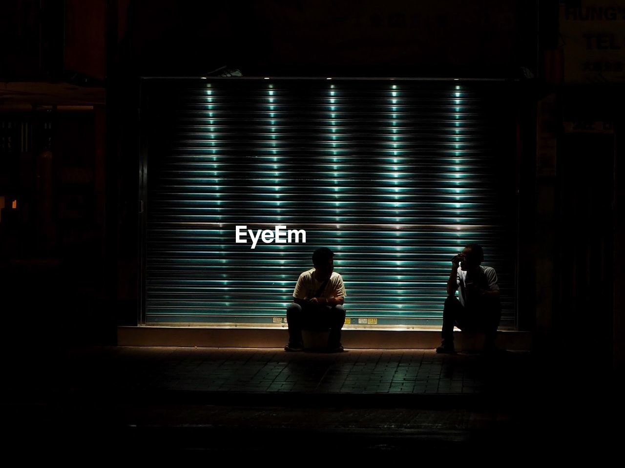 Men sitting against illuminated shutter in city at night