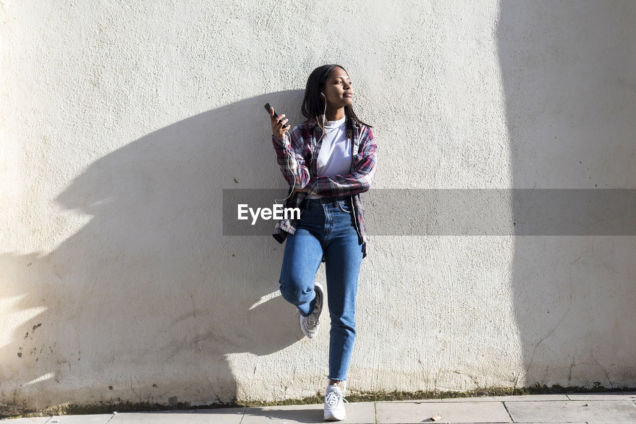 Young woman listening to music while leaning against wall