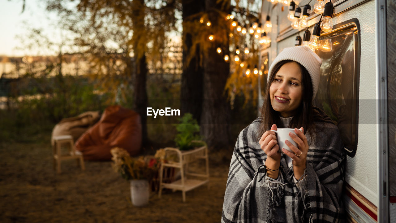 Caucasian woman in a knitted hat wrapped in a plaid and drinks a warming drink outdoors. 