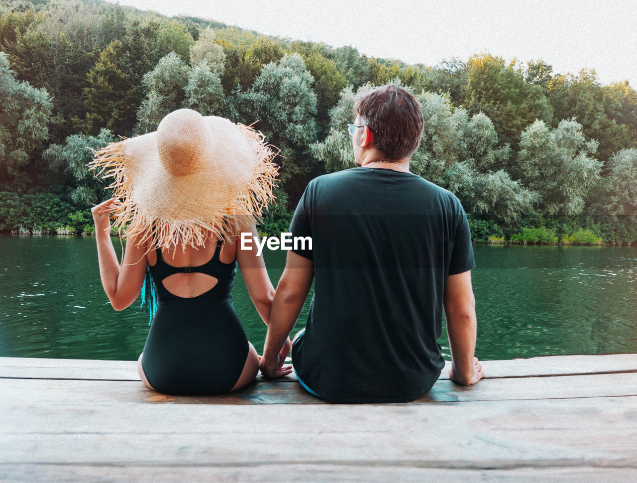 Rear view of couple sitting on pier in lake against trees