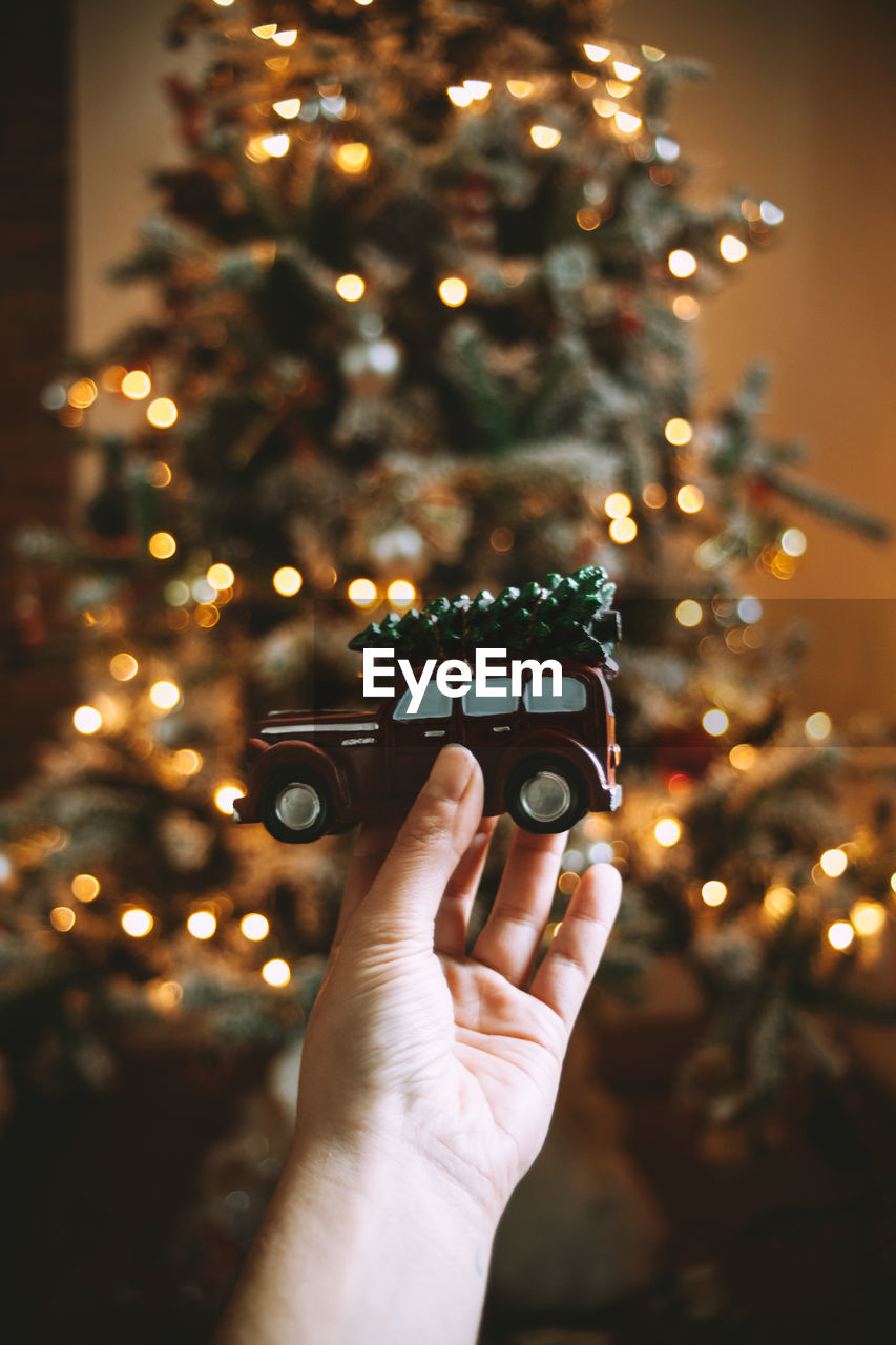 Cropped hand of woman holding toy car against illuminated christmas tree