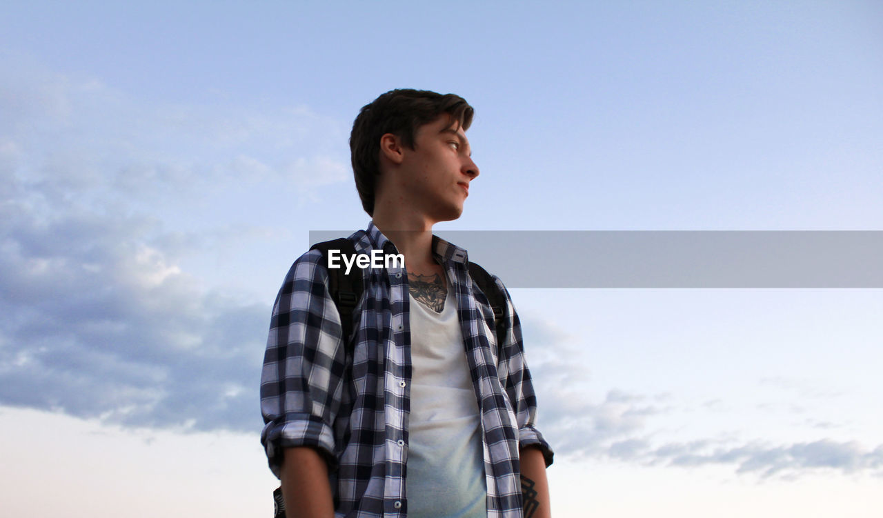 Low angle view of young man looking away while standing against sky