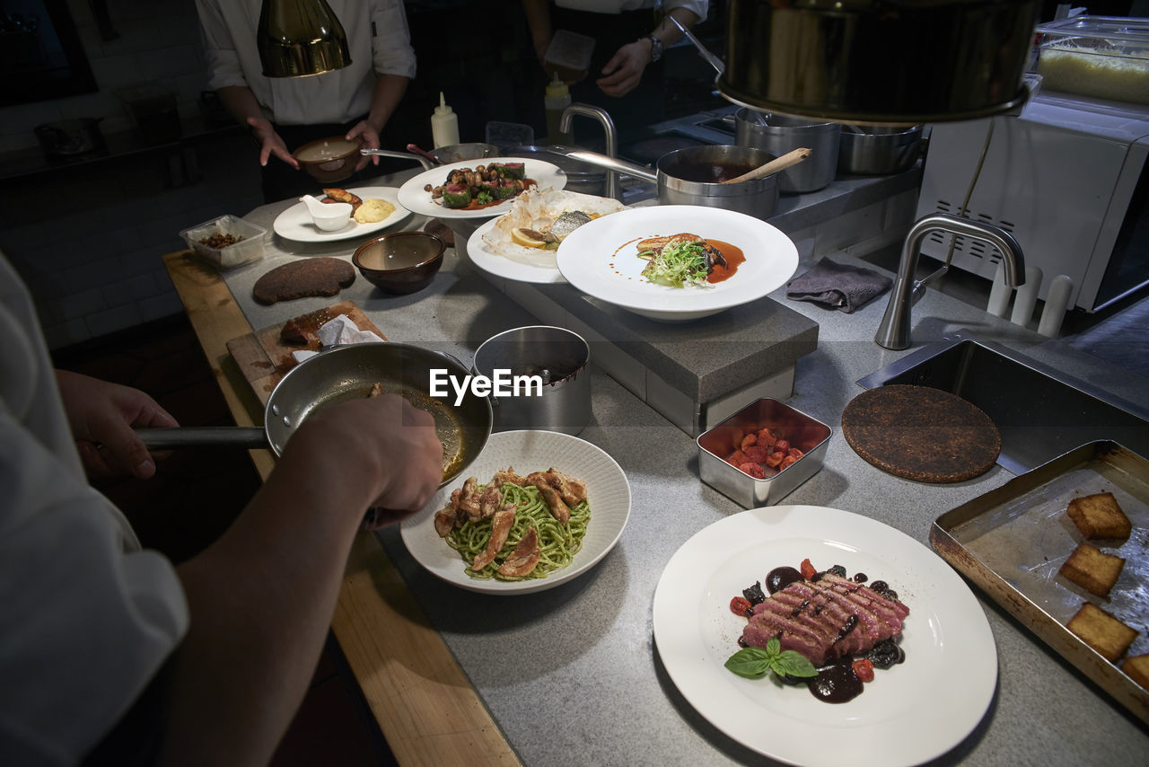 High angle of crop anonymous chef adding fried chicken fillets into plate with pasta while preparing food with colleagues in modern restaurant kitchen