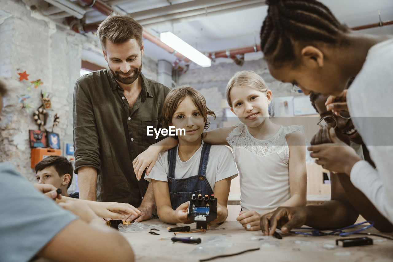 Portrait of smiling girl with arm around female friend amidst teacher and pupils at technology workshop in school