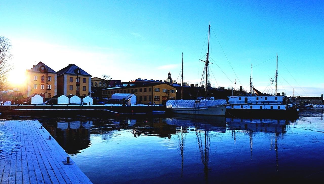 Sailboats moored in river in front of houses against blue sky during winter