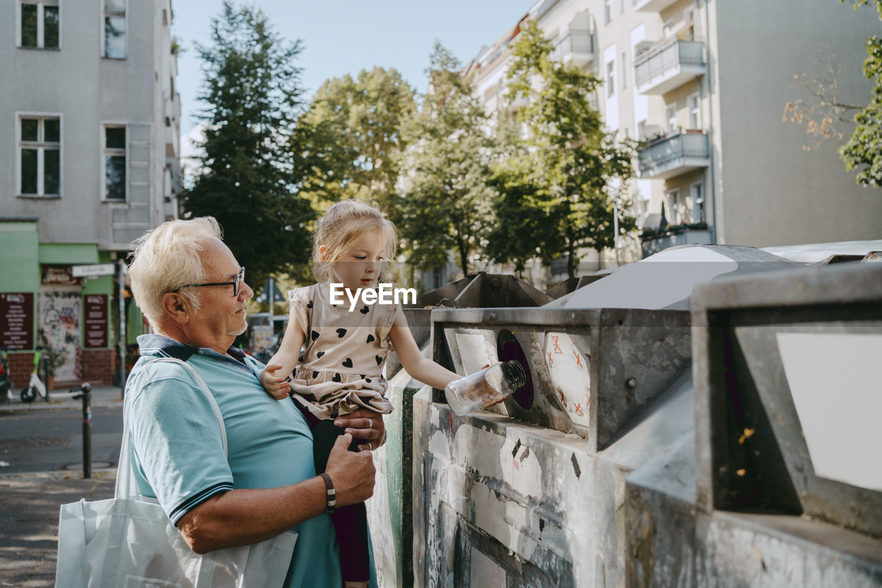 Senior man carrying granddaughter trashing garbage in can at street