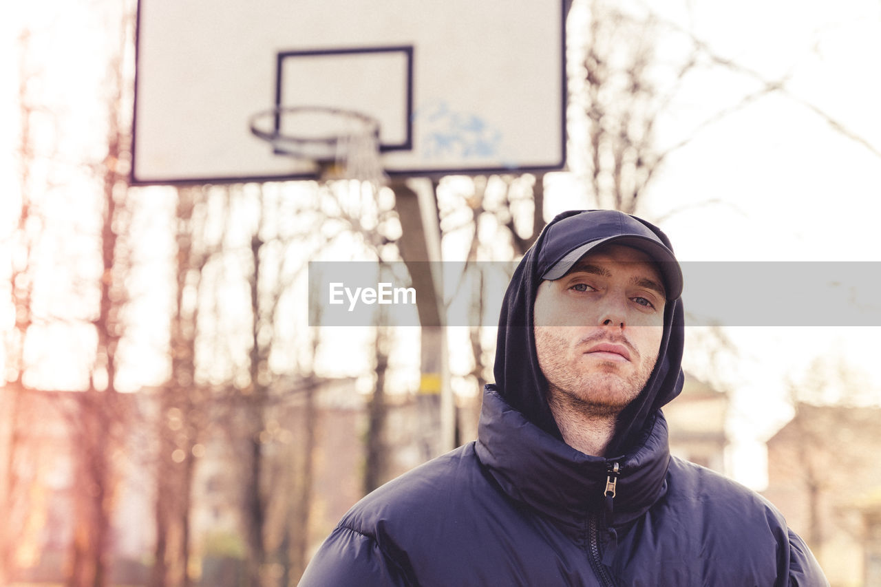 Portrait of young man standing against basketball hoop and trees