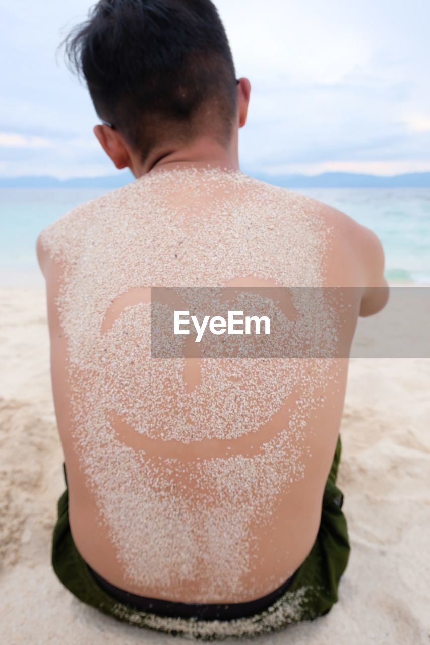 Rear view of shirtless man with anthropomorphic smiley face on his back at beach against sky