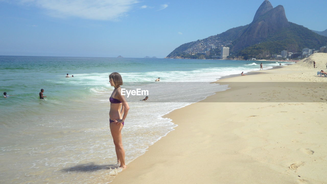 Young woman in bikini standing at shore against sky