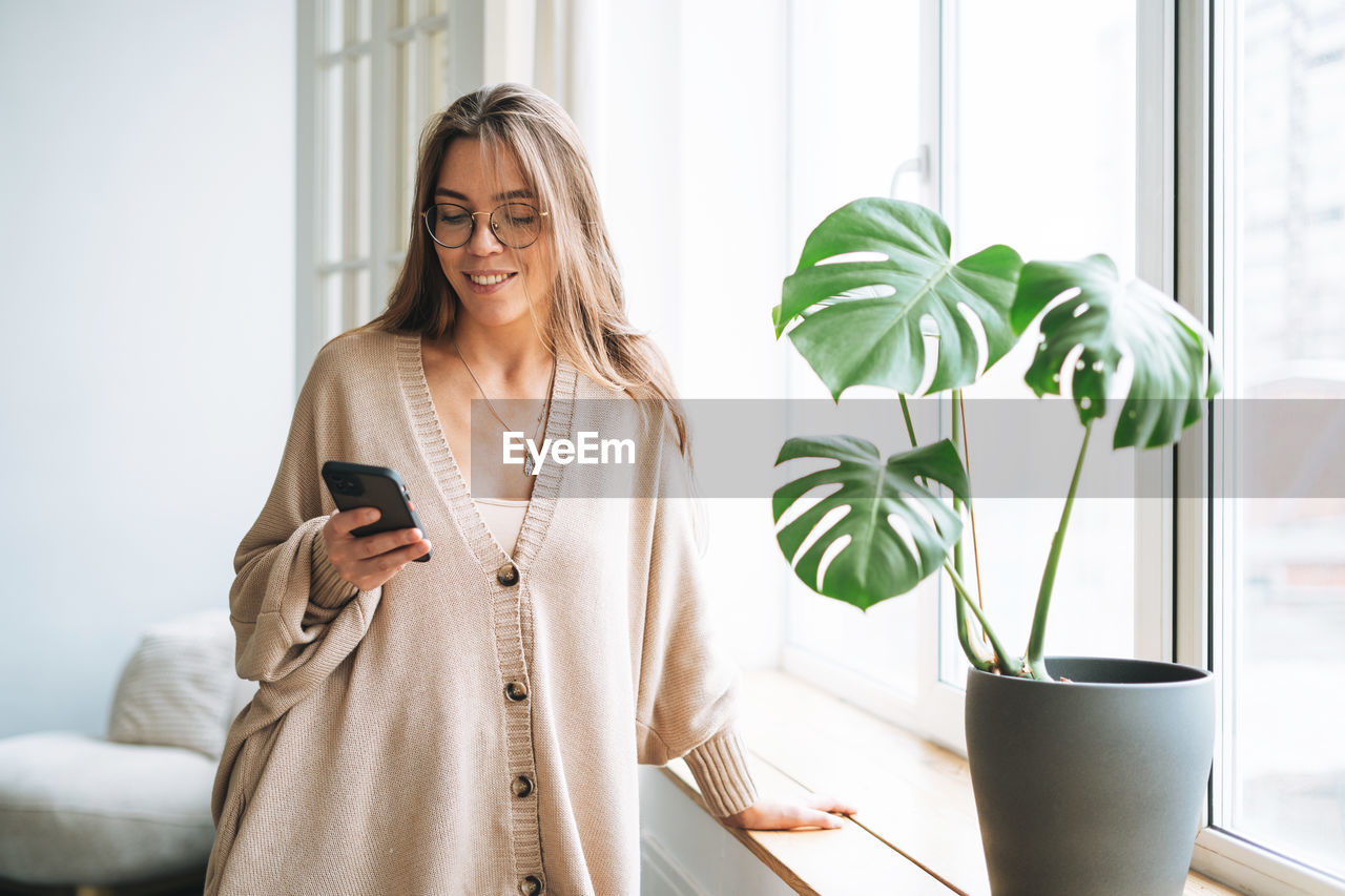 Young beautiful woman with in cozy knitted cardigan using mobile phone in bright interior at home