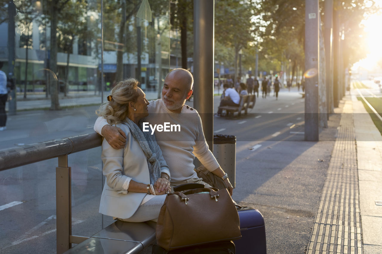 Spain, barcelona, senior couple with baggage sitting at tram stop in the city at sunset