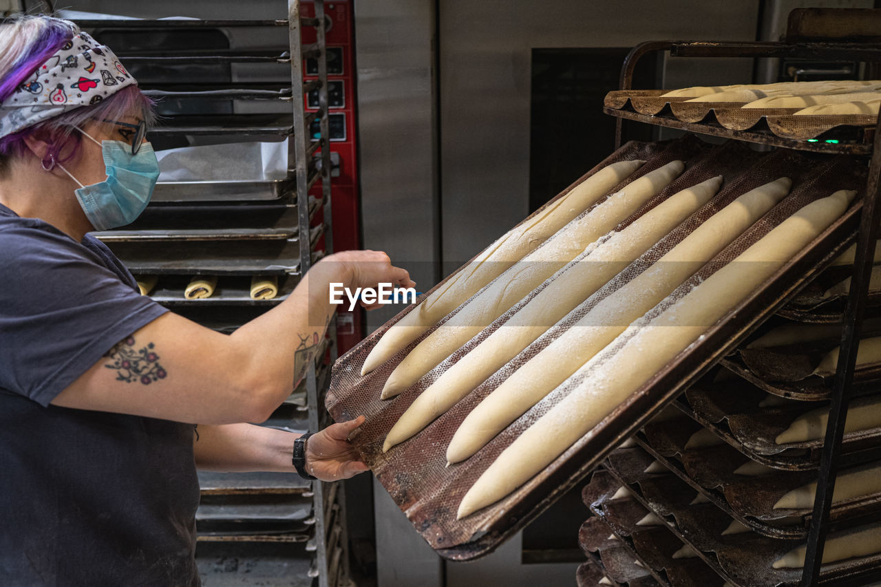 Side view of focused female baker in protective mask making notches on raw baguettes placed on baking pan in bakehouse