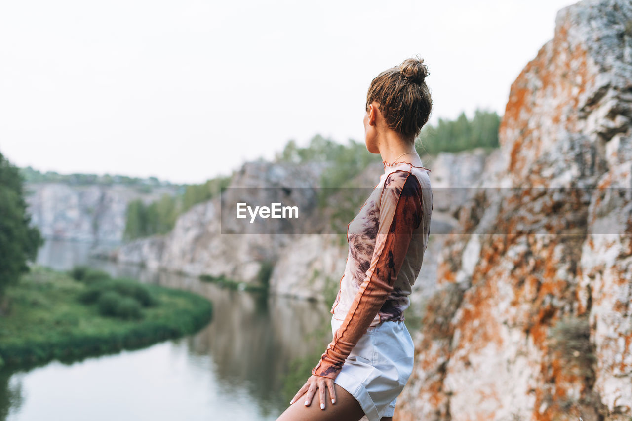 Young slim woman in casual clothes looks at beautiful view of mountains and calm river, local travel