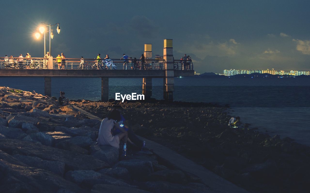 People sitting on rock next to jetty against cloudy sky