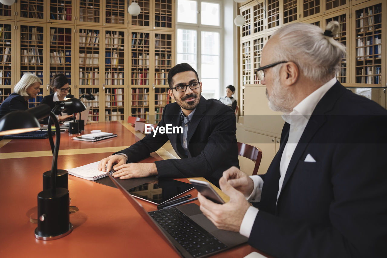 Smiling man discussing with senior lawyer at table in library