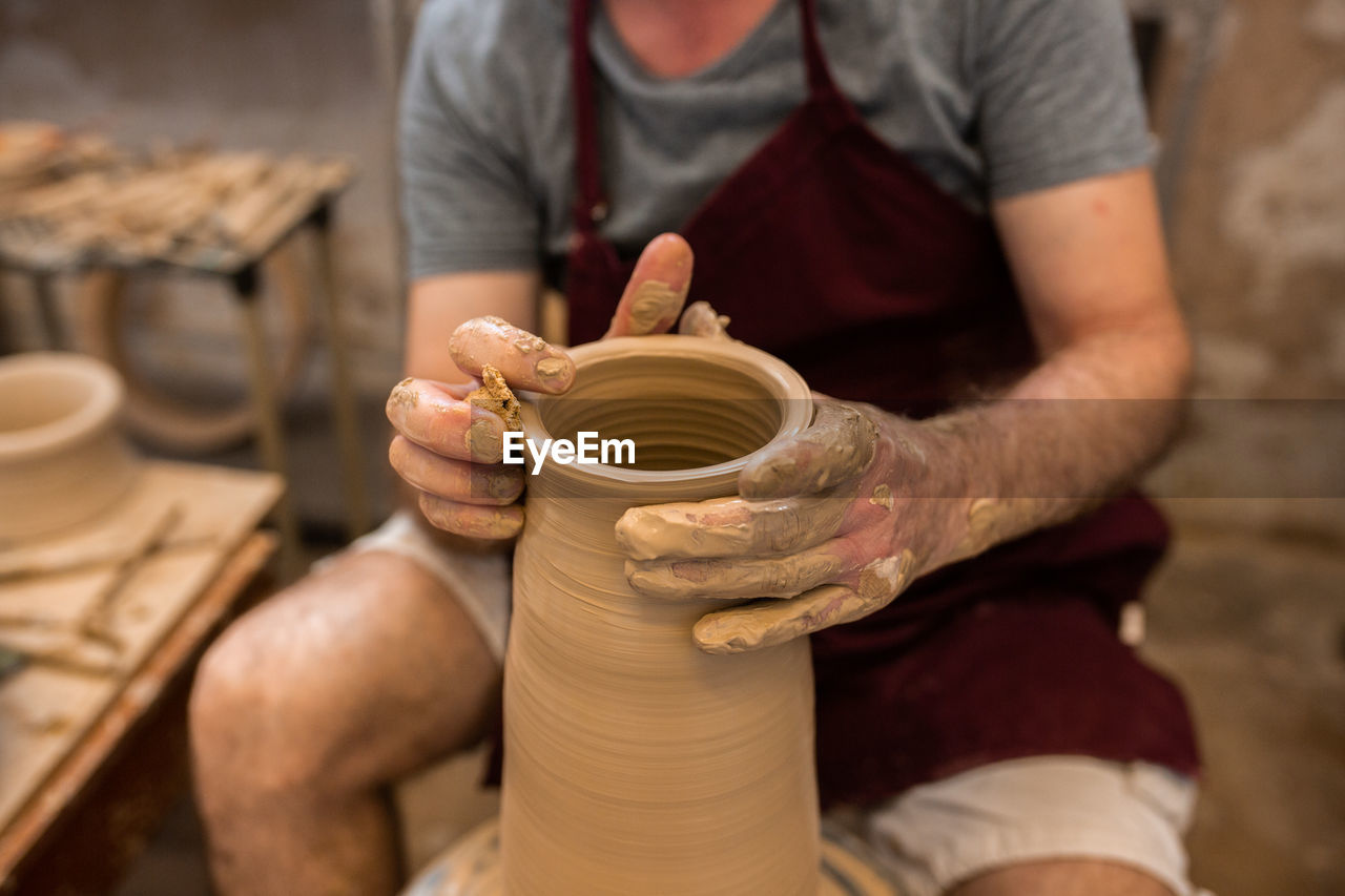 Crop unrecognizable sculptor with equipment giving shape while sculpting with brown clay on throwing wheel