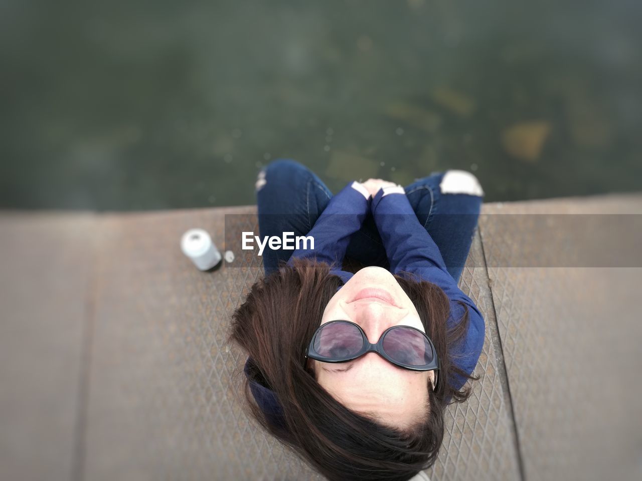 High angle portrait of smiling young woman in sunglasses sitting on jetty over lake