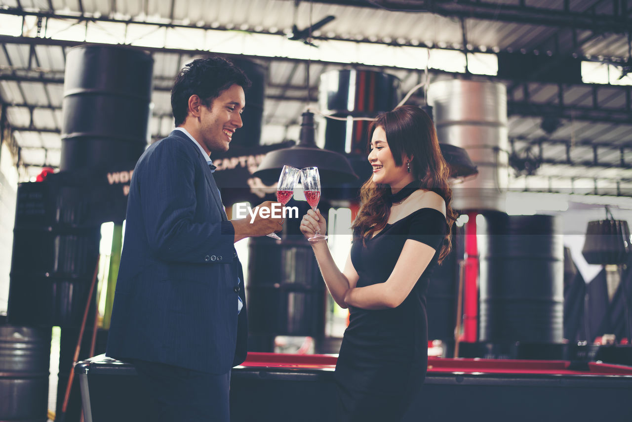 Side view of smiling couple toasting red wineglasses while standing in bar