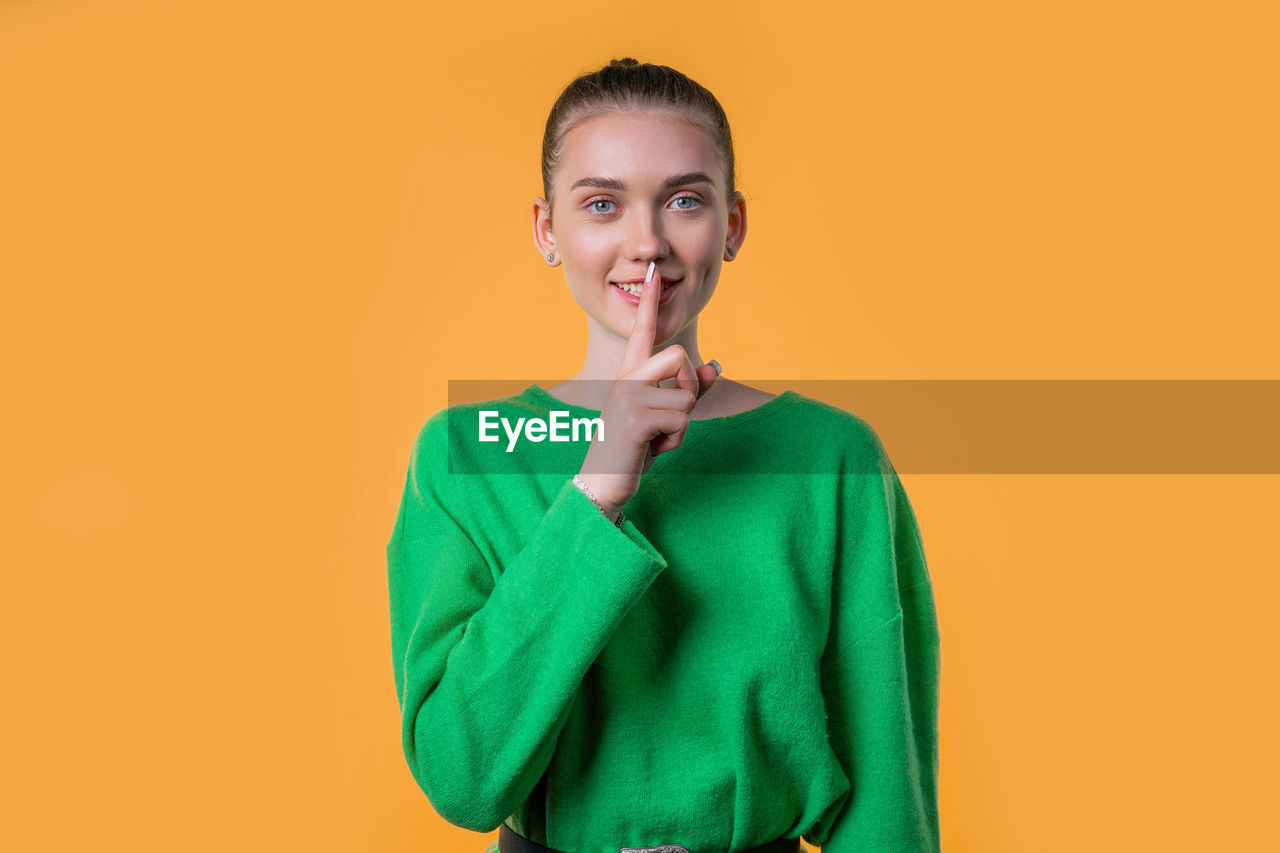 studio shot, portrait, one person, looking at camera, smiling, colored background, adult, indoors, happiness, emotion, women, young adult, outerwear, copy space, waist up, clothing, front view, standing, sleeve, casual clothing, green, cheerful, orange color, yellow, sweater, smile, teeth, positive emotion, female, cut out, person, sweatshirt, hairstyle, brown hair