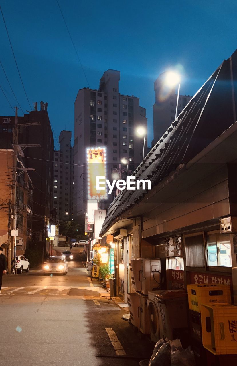 ILLUMINATED ROAD AMIDST BUILDINGS AGAINST SKY AT NIGHT