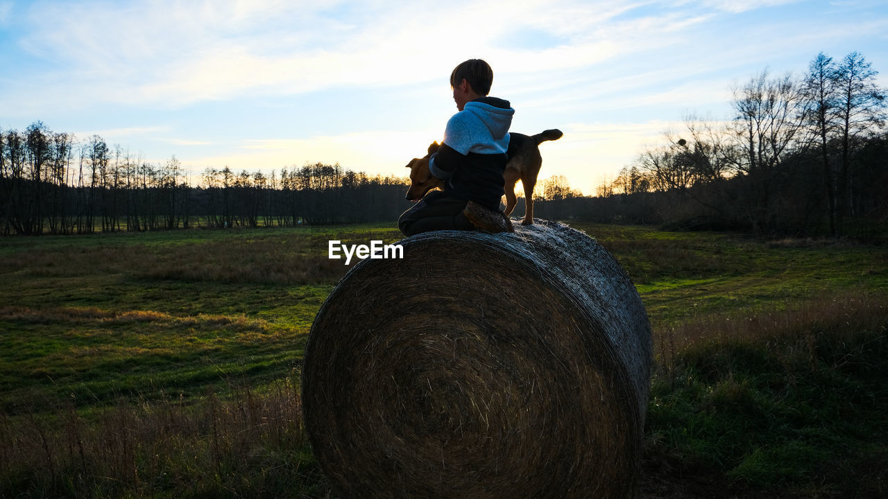 Boy and dog sitting on bale of straße in front of field against sky