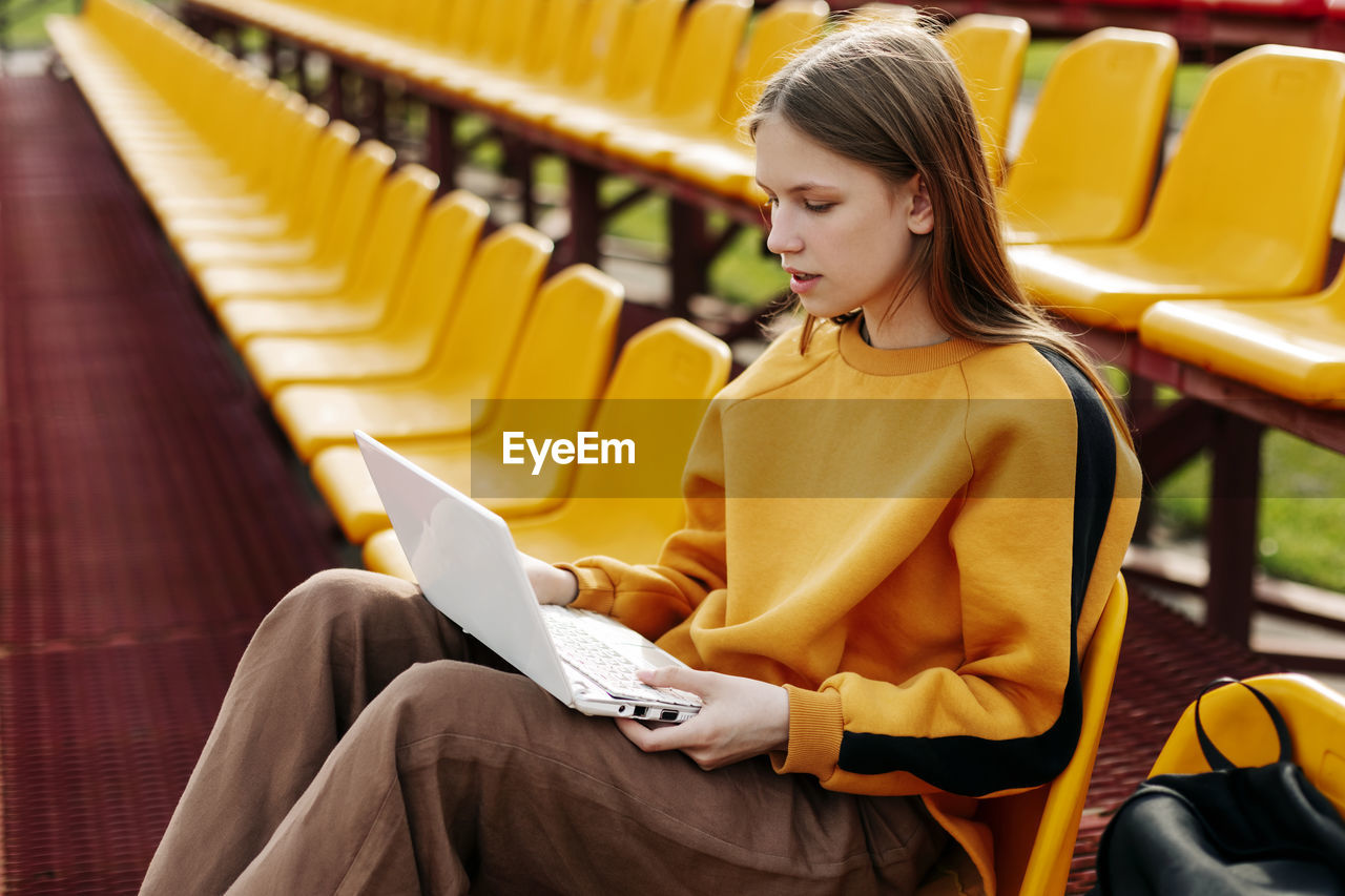 A young girl is doing her homework using a laptop in the stands of the stadium. 