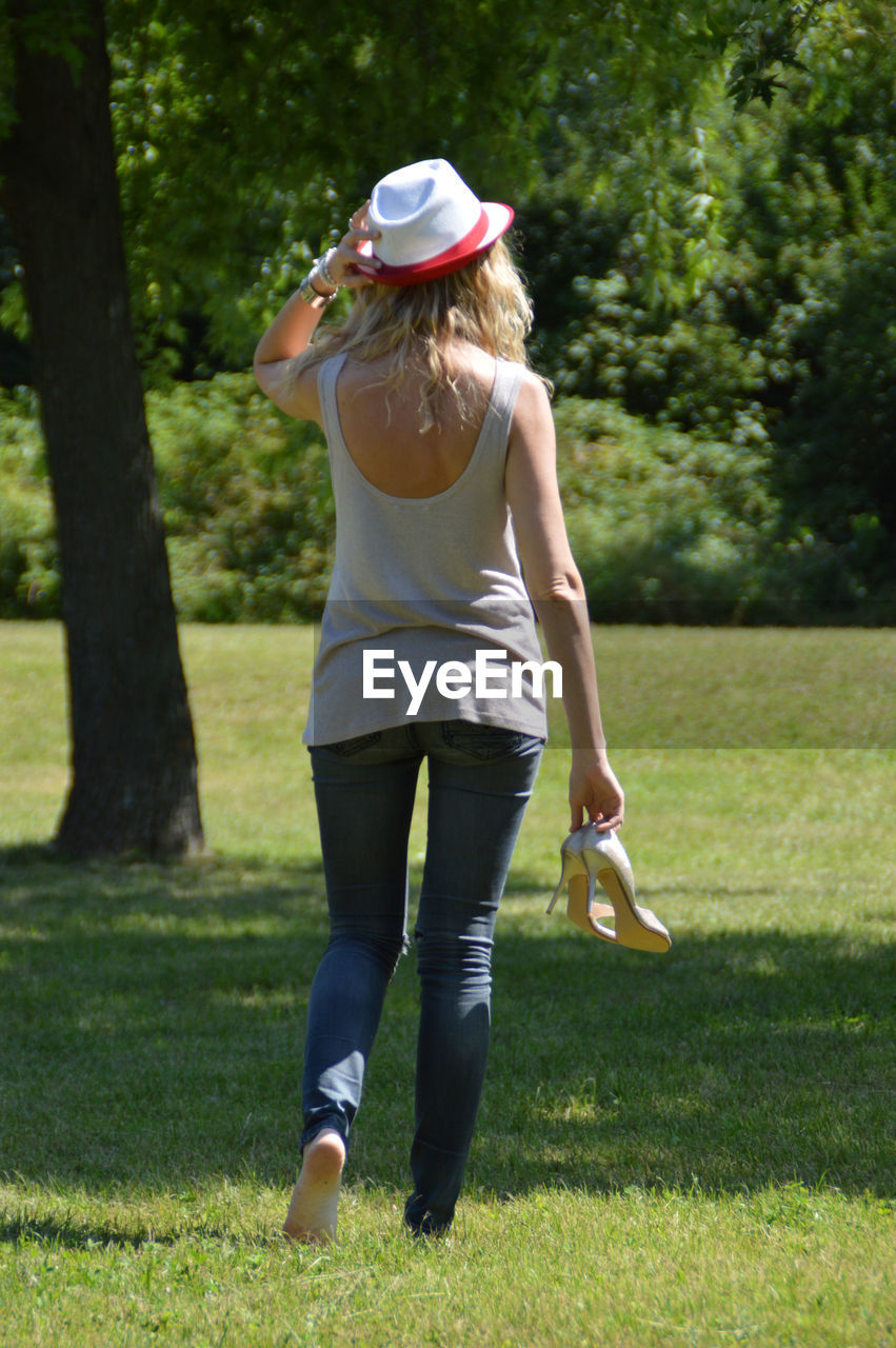 Rear view of woman holding high heels walking on grass at park