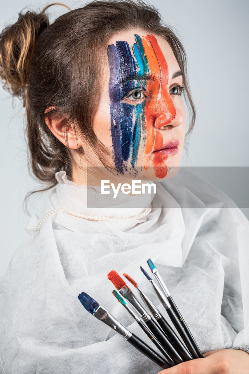 Young woman with painted face holding paintbrushes against gray background