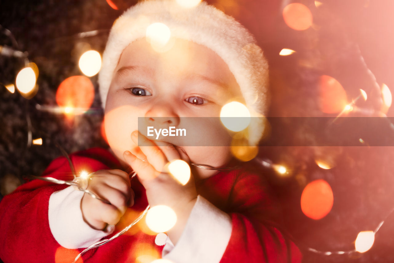 Close-up of cute baby boy in santa costume with illuminated string lights