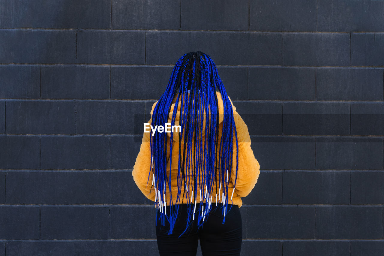 Back view of unrecognizable female with blue braided hairstyle and in yellow coat standing in street