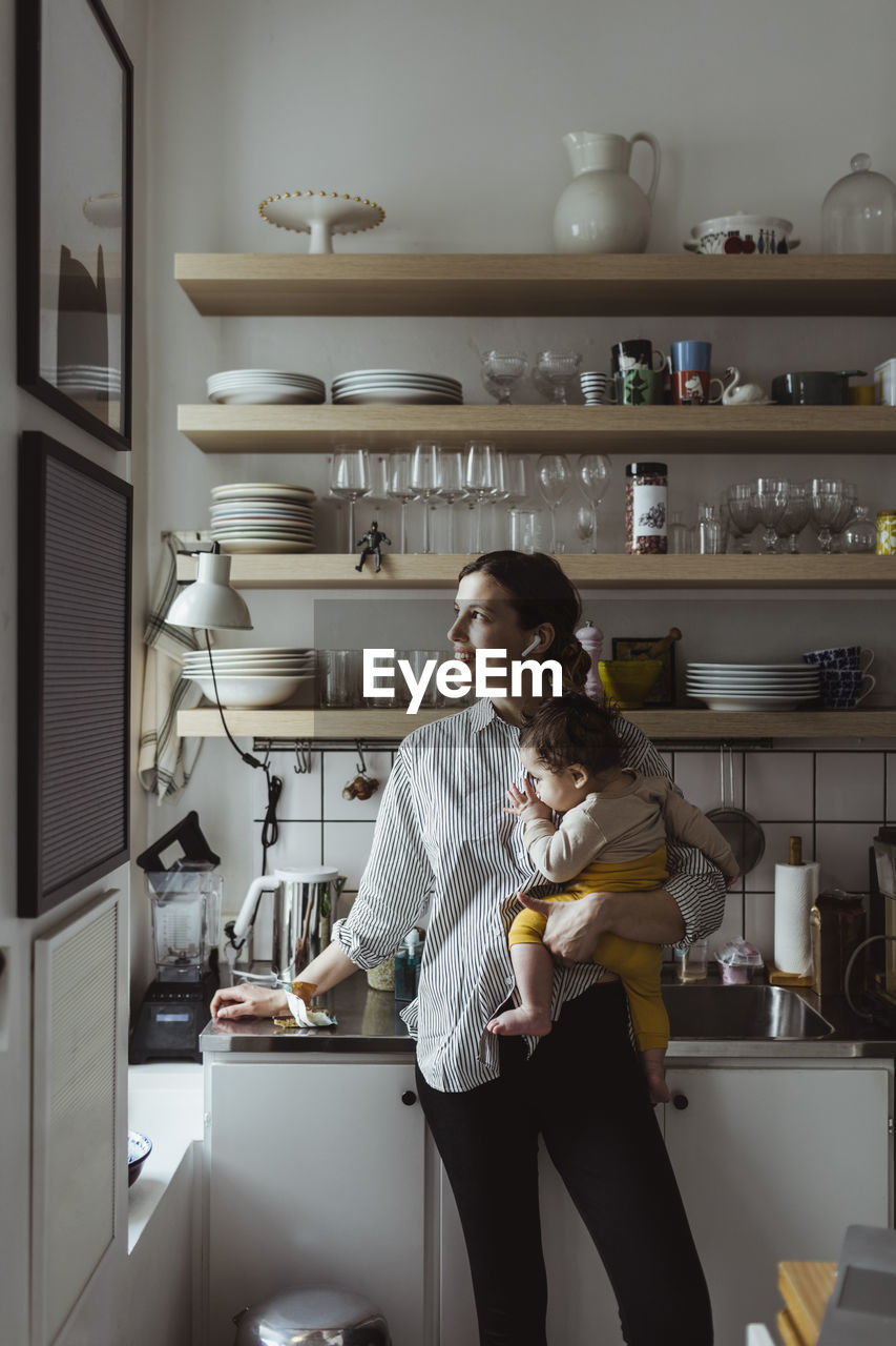 Smiling mother looking away while carrying male toddler in kitchen
