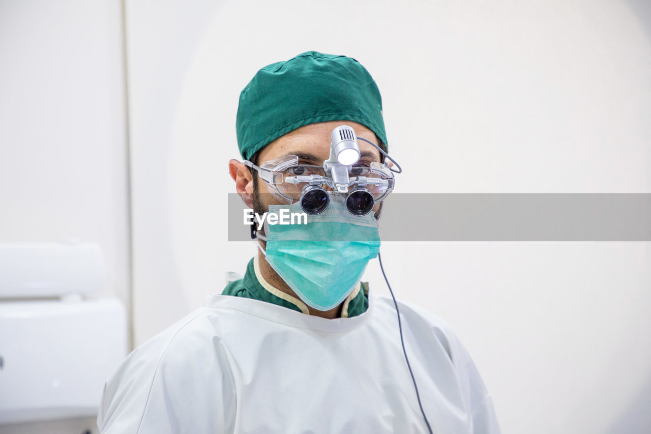 Male doctor in medical mask and bilocular glasses standing in modern dental clinic and looking at camera