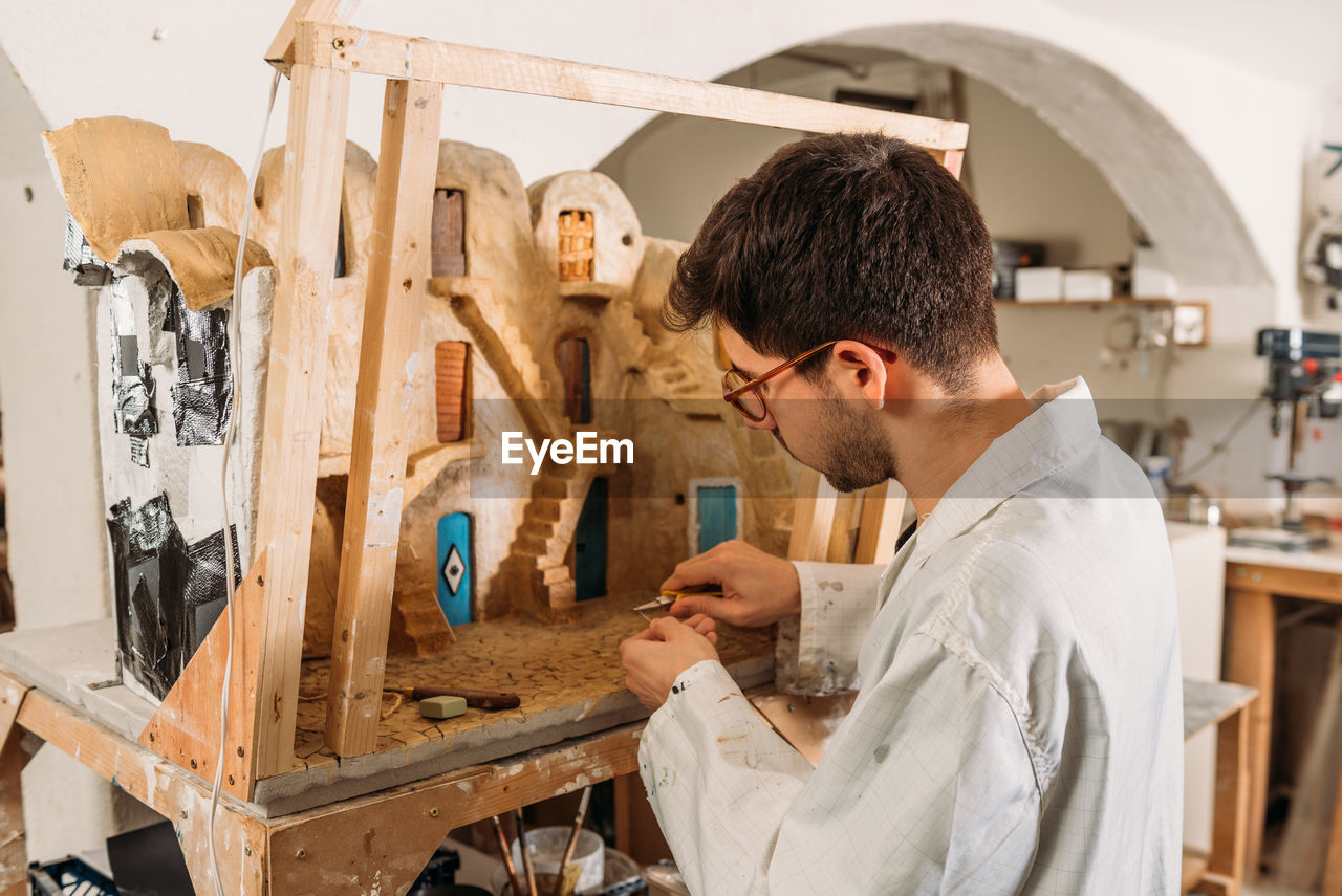 Side view of craftsman making model of facade of clay house and creating small wooden door while working in art workshop