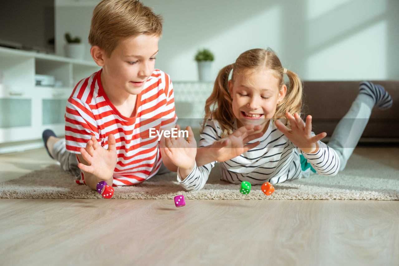 Cheerful siblings playing with dice while lying on carpet at home