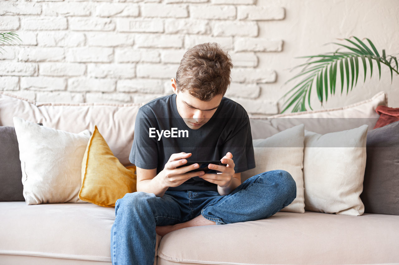 young man using mobile phone while sitting on sofa against wall