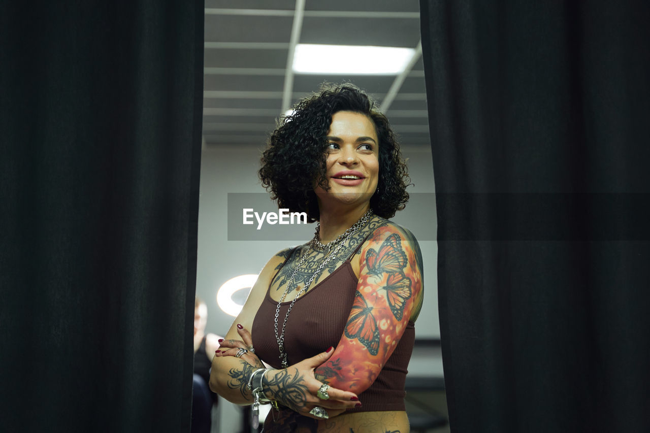 Positive adult woman in casual clothes with tattoos standing in light tattoo salon while looking away with crossed hands