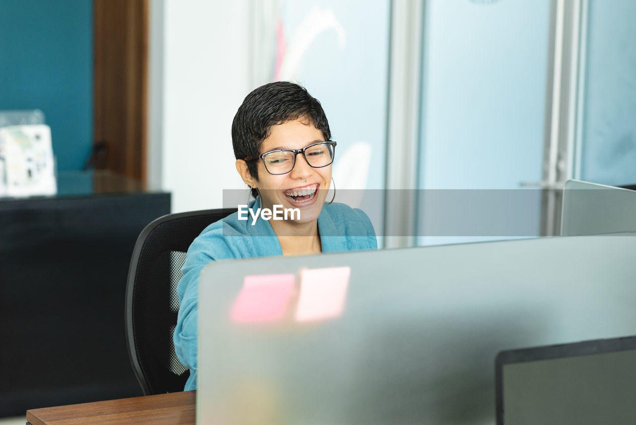 Cheerful hispanic female entrepreneur with short hair in stylish outfit sitting at table with laptop and laughing while working in light office in costa rica in daytime