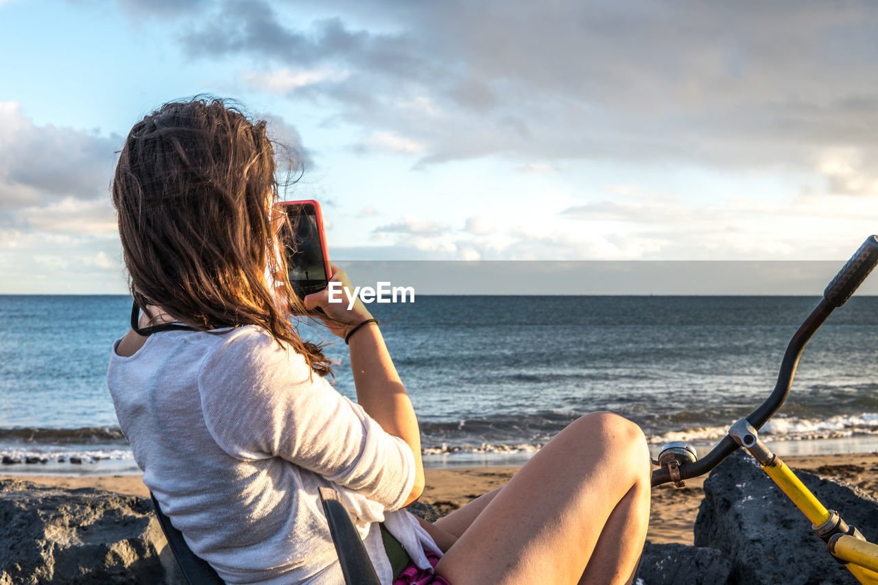 Side view of woman photographing sea with mobile phone against sky during sunset