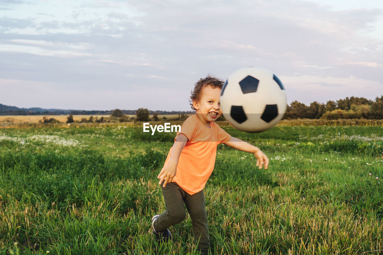 Smiling boy playing with soccer ball on grassy land against sky