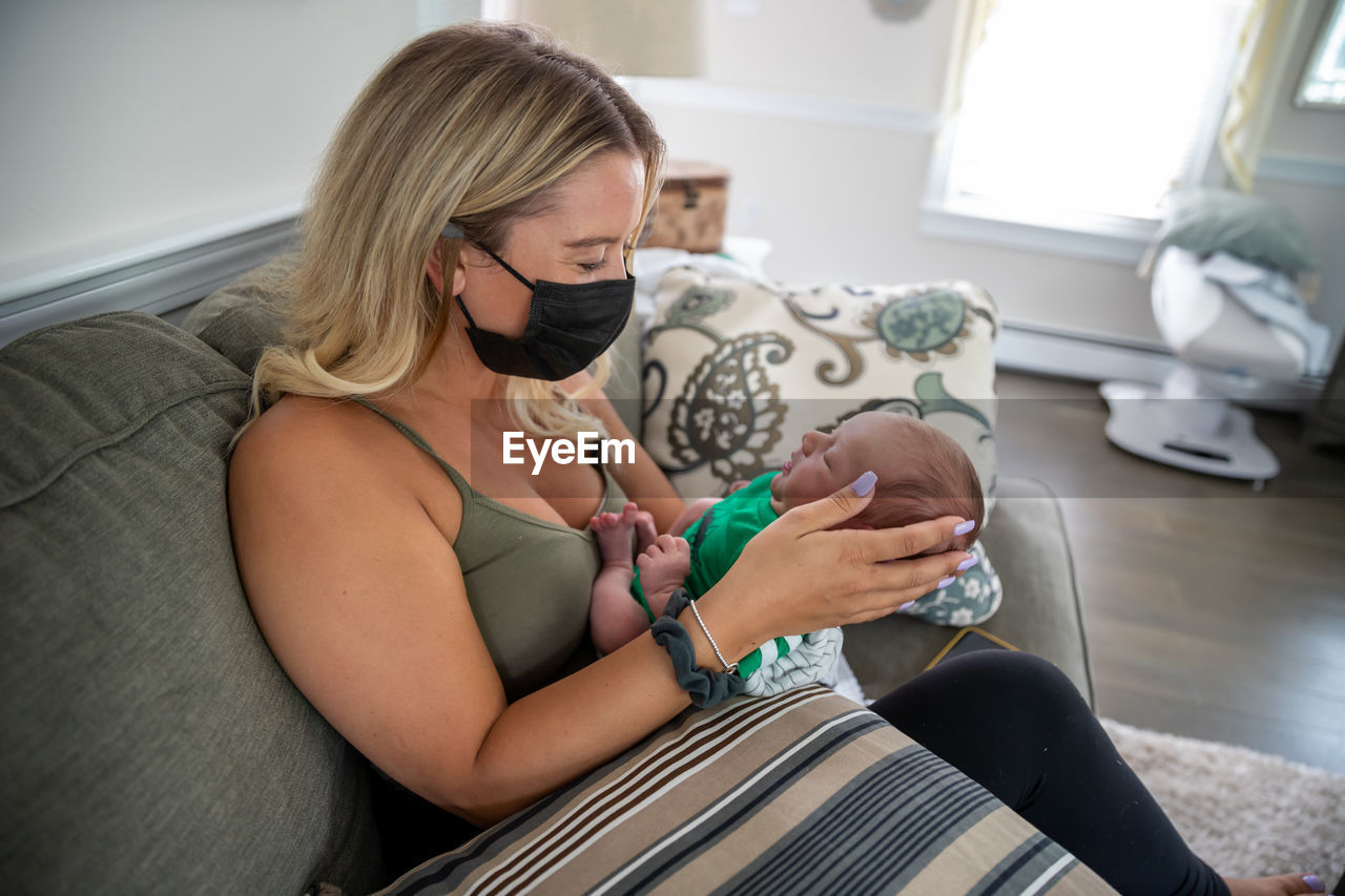 Blonde woman wearing face mask holding sleepy newborn baby.