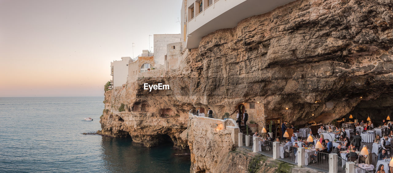 Panoramic view of people at restaurant amidst rocks by sea against clear sky