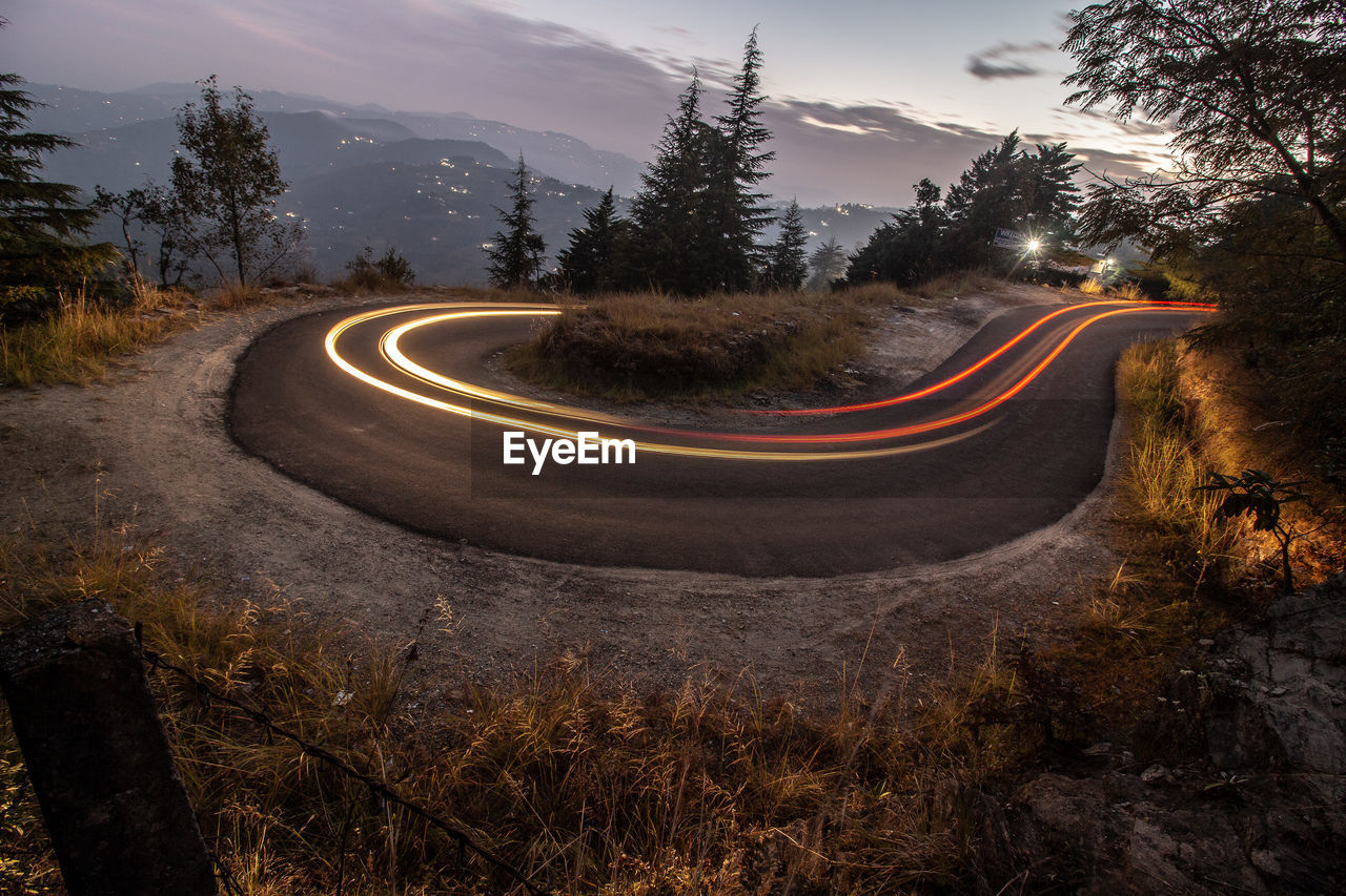 Light trails on road by trees against sky in city