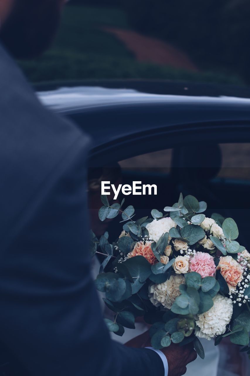 Cropped hand of bridegroom holding bouquet by car
